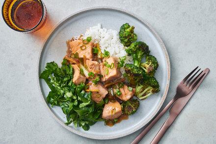 مکعبهای سالمون با لعاب سبک سویا-زنجبیل پوشانده شده و با پیازچه برشخورده تزیین شدهاند. آنها روی تخت برنج با بروکلی سرخشده در یک سمت و سالاد آرگولا در سمت دیگر قرار دارند؛ یک نوشیدنی و چاقو و چنگال در نزدیکی قرار دارند.